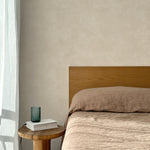 Bed with tan linens and wooden headboard, wooden side table  with white book and green glass, white floor length curtain against a white celestial dotted pattern on a soft beige background. 