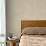 Bed with tan linens and wooden headboard, wooden side table  with white book and green glass, white floor length curtain against a white celestial dotted pattern on a soft beige background. 