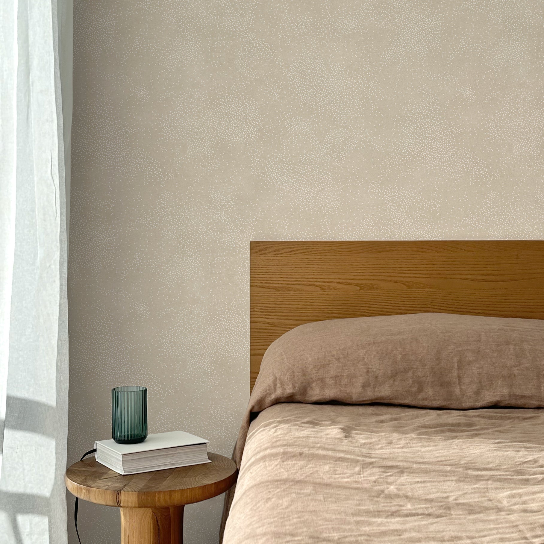 Bed with tan linens and wooden headboard, wooden side table  with white book and green glass, white floor length curtain against a white celestial dotted pattern on a soft beige background. 