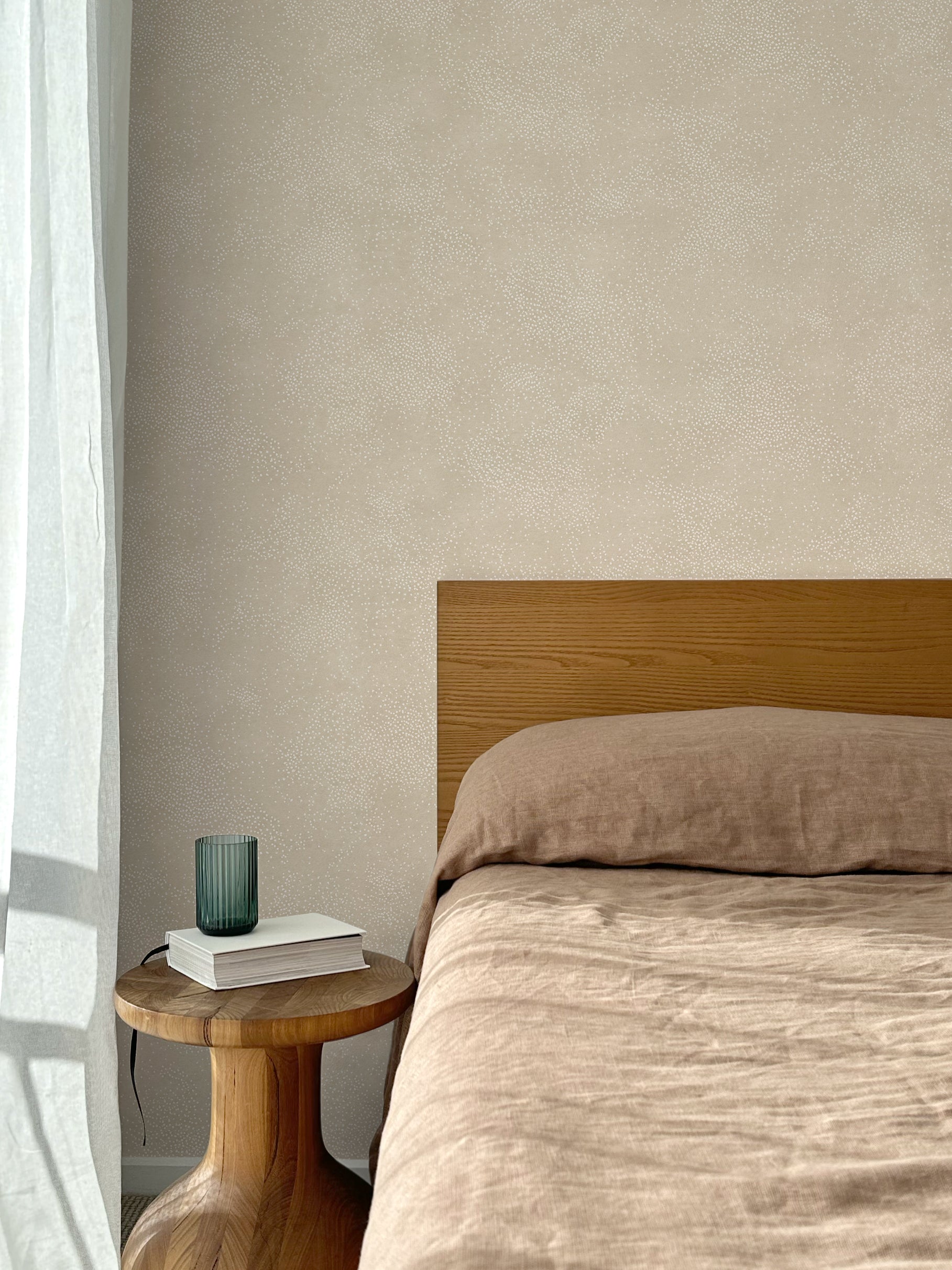 Bed with tan linens and wooden headboard, wooden side table  with white book and green glass, white floor length curtain against a white celestial dotted pattern on a soft beige background. 