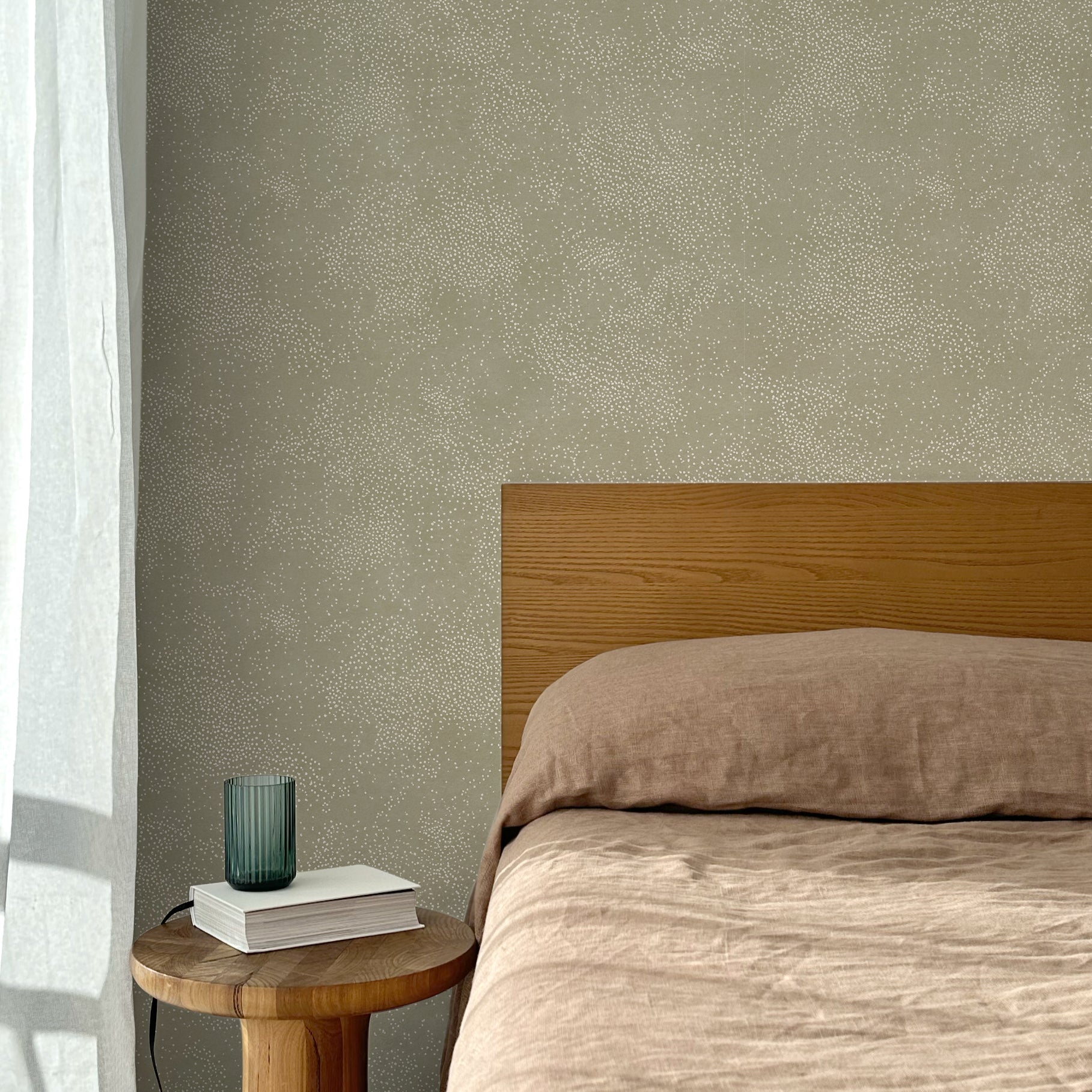 Bed with tan linens and wooden headboard, wooden side table  with white book and green glass, white floor length curtain against a white celestial dotted pattern on a sage green background. 