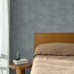 Bed with tan linens and wooden headboard, wooden side table  with white book and green glass, white floor length curtain against a white celestial dotted pattern on a cool blue background. 