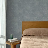Bed with tan linens and wooden headboard, wooden side table  with white book and green glass, white floor length curtain against a white celestial dotted pattern on a cool blue background. 