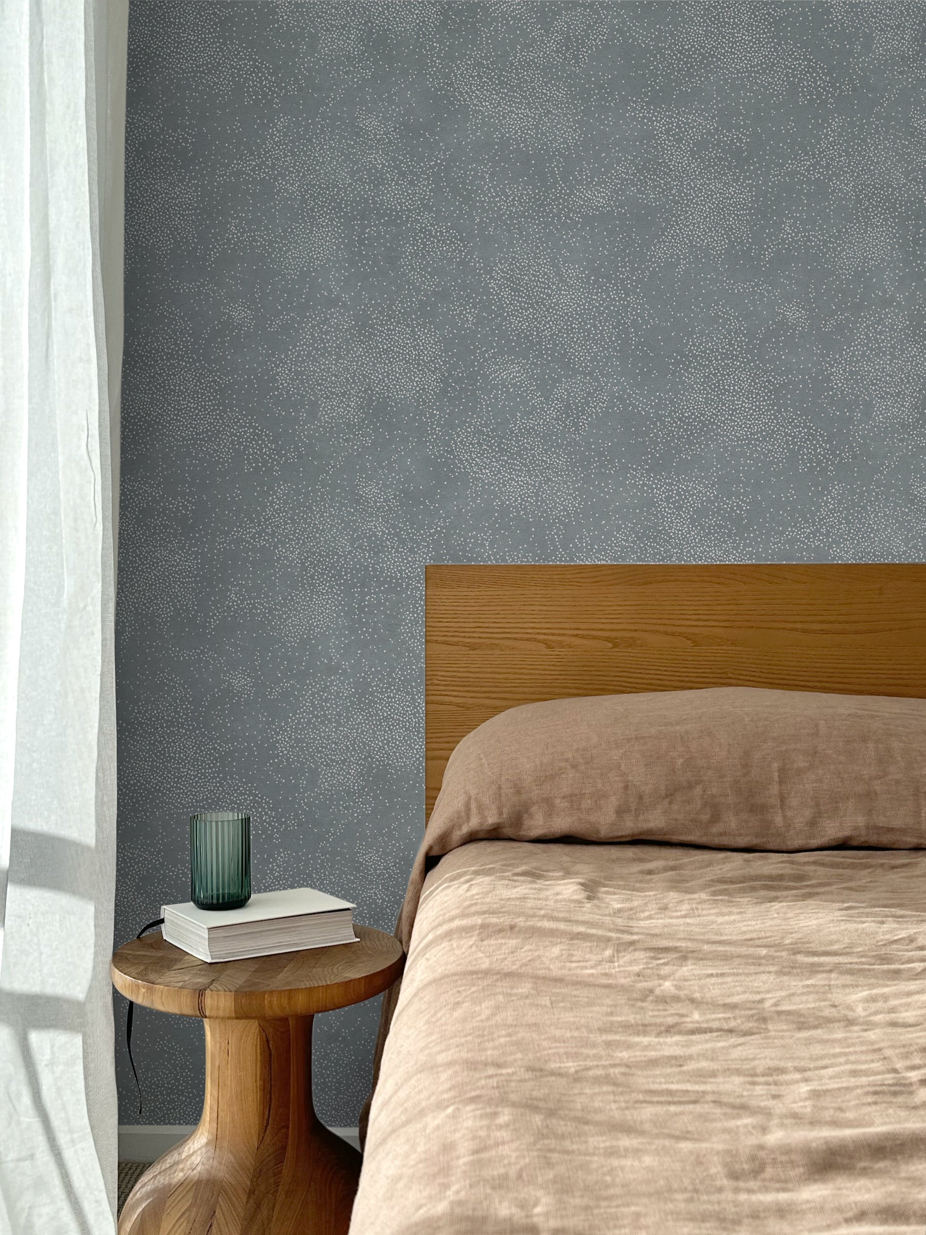 Bed with tan linens and wooden headboard, wooden side table  with white book and green glass, white floor length curtain against a white celestial dotted pattern on a cool blue background. 