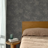 Bed with tan linens and wooden headboard, wooden side table  with white book and green glass, white floor length curtain against a white celestial dotted pattern on a charcoal grey background. 