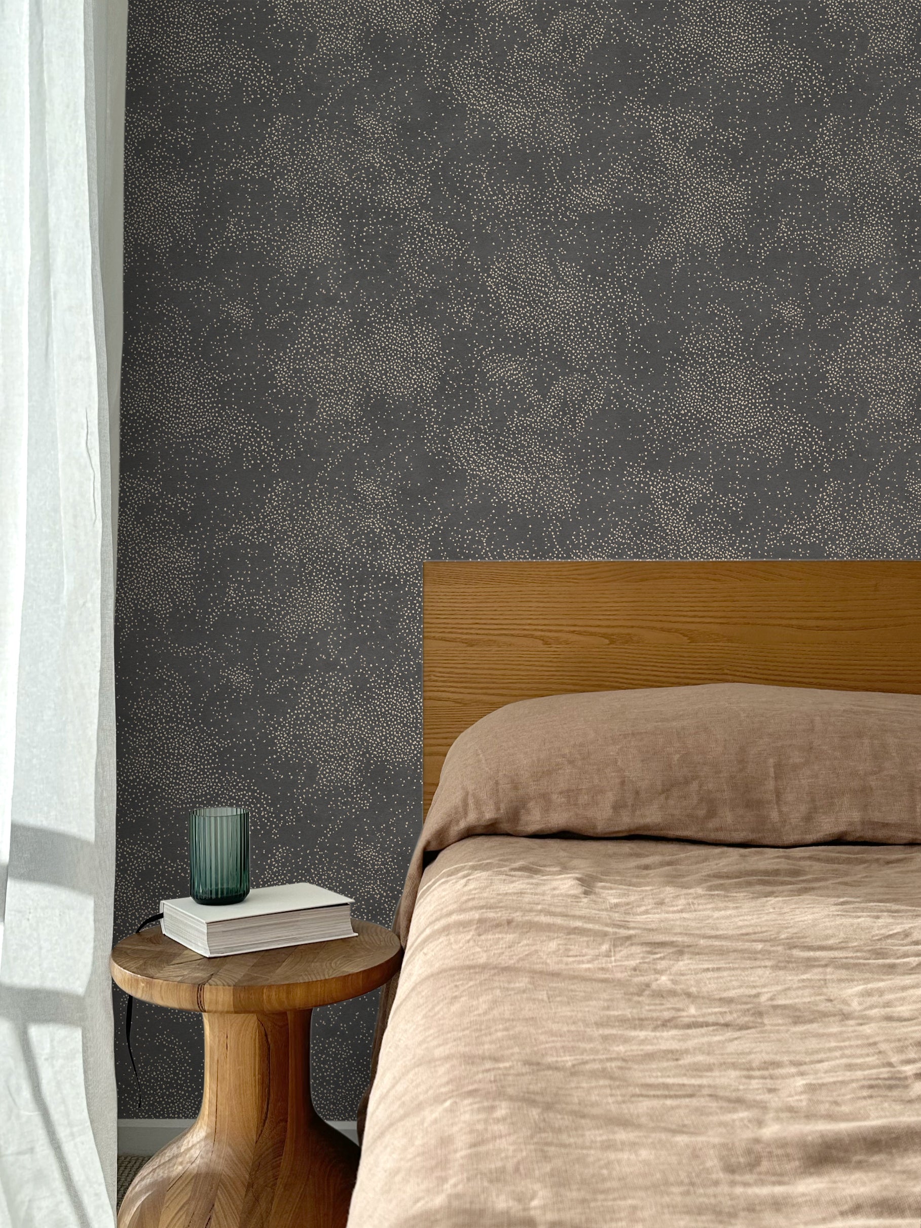 Bed with tan linens and wooden headboard, wooden side table  with white book and green glass, white floor length curtain against a white celestial dotted pattern on a charcoal grey background. 