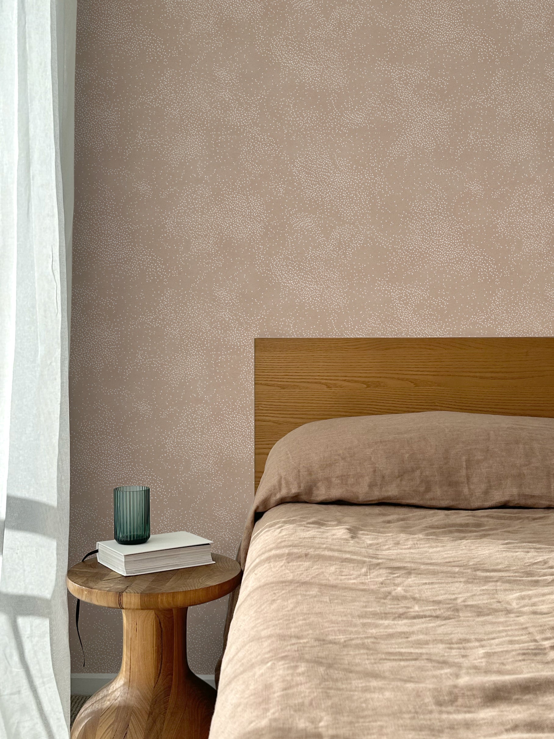 Bed with tan linens and wooden headboard, wooden side table  with white book and green glass, white floor length curtain against a white celestial dotted pattern on a clay pink background.