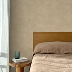Bed with tan linens and wooden headboard, wooden side table  with white book and green glass, white floor length curtain against a white celestial dotted pattern on a tan background. 