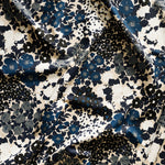 Close up of gently draped printed linen fabric with a dense flower and leaf pattern in shades of dark blue, grey, white and black on a beige background.