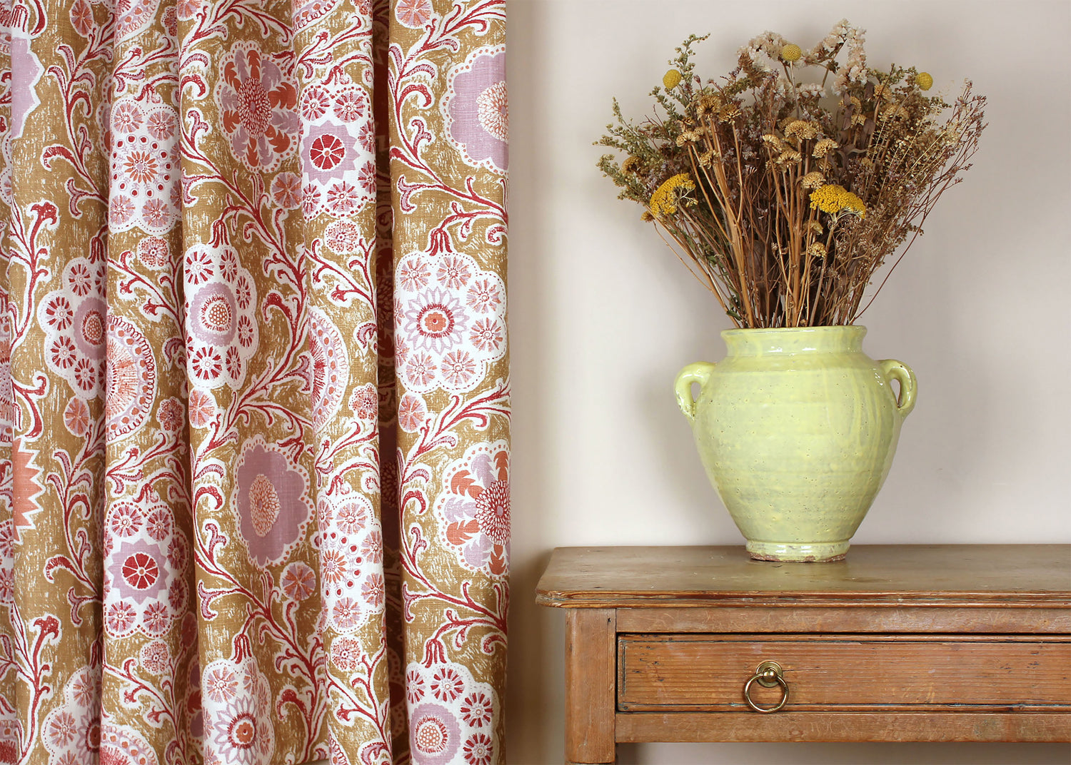 Curtain in a handdrawn playful floral design in coral, lilac and red against a tan field along a side a wooden table with vase and dried flowers. 