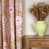 Curtain in a handdrawn playful floral design in coral, lilac and red against a tan field along a side a wooden table with vase and dried flowers. 
