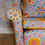 Chair upholstered in printed linen in a handdrawn playful floral design in red, yellow, green and blue on a light blue field against matching wallpaper.