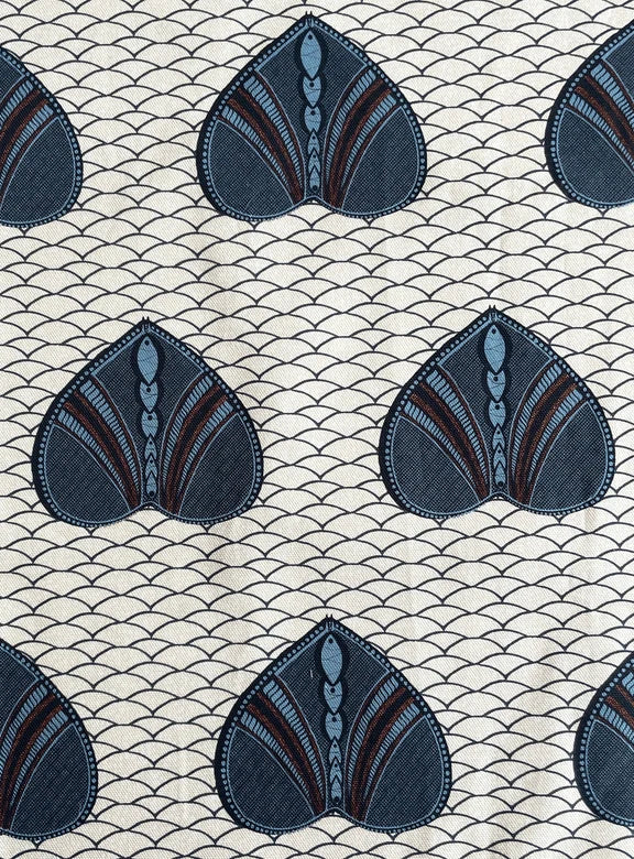 A close-up of a fabric pattern featuring repeating stylized fan-shaped motifs in dark blue, slate blue, and brown on a cream background with a fine repeating wave line pattern.