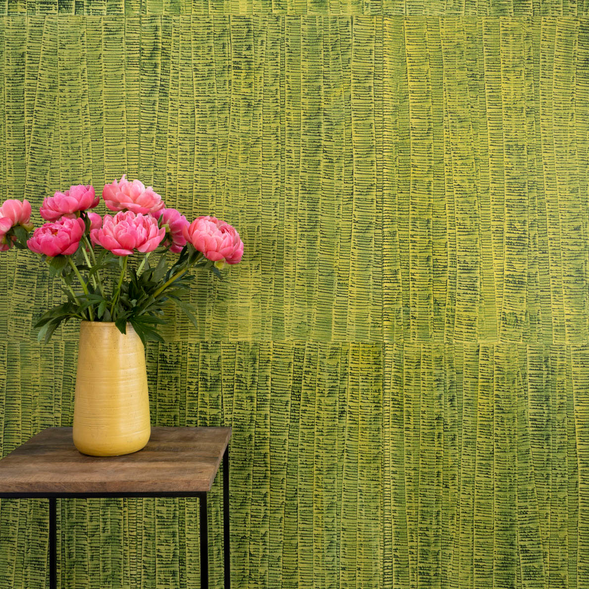 A vase of flowers stands in front of a wall papered in an undulating ribbon pattern in green on a gold field.