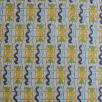 Close up of a printed fabric with blue squiggles, yellow striped triangles,  yellow X marks on a broken grid in green on a white background. 