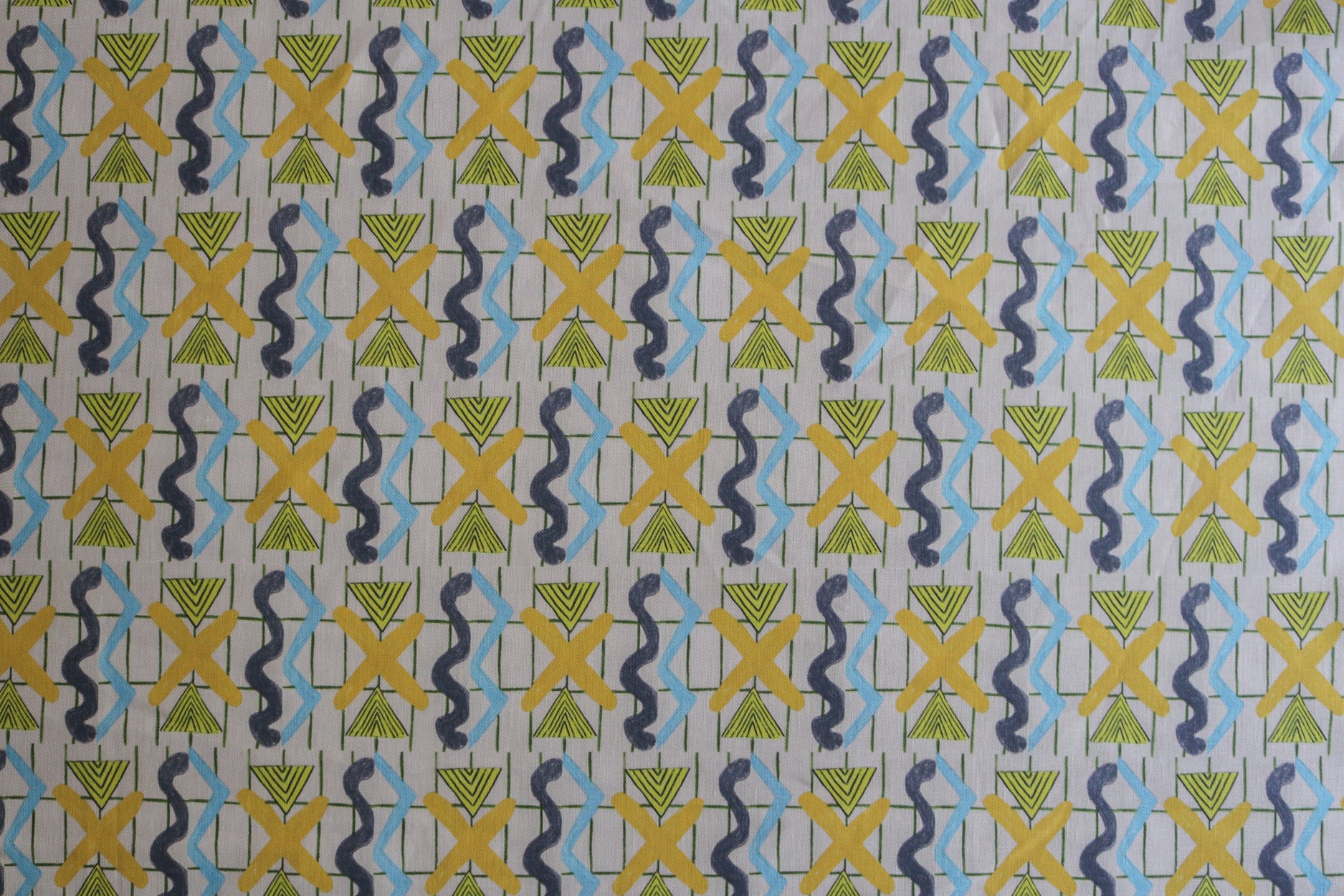 Close up of a printed fabric with blue squiggles, yellow striped triangles,  yellow X marks on a broken grid in green on a white background. 