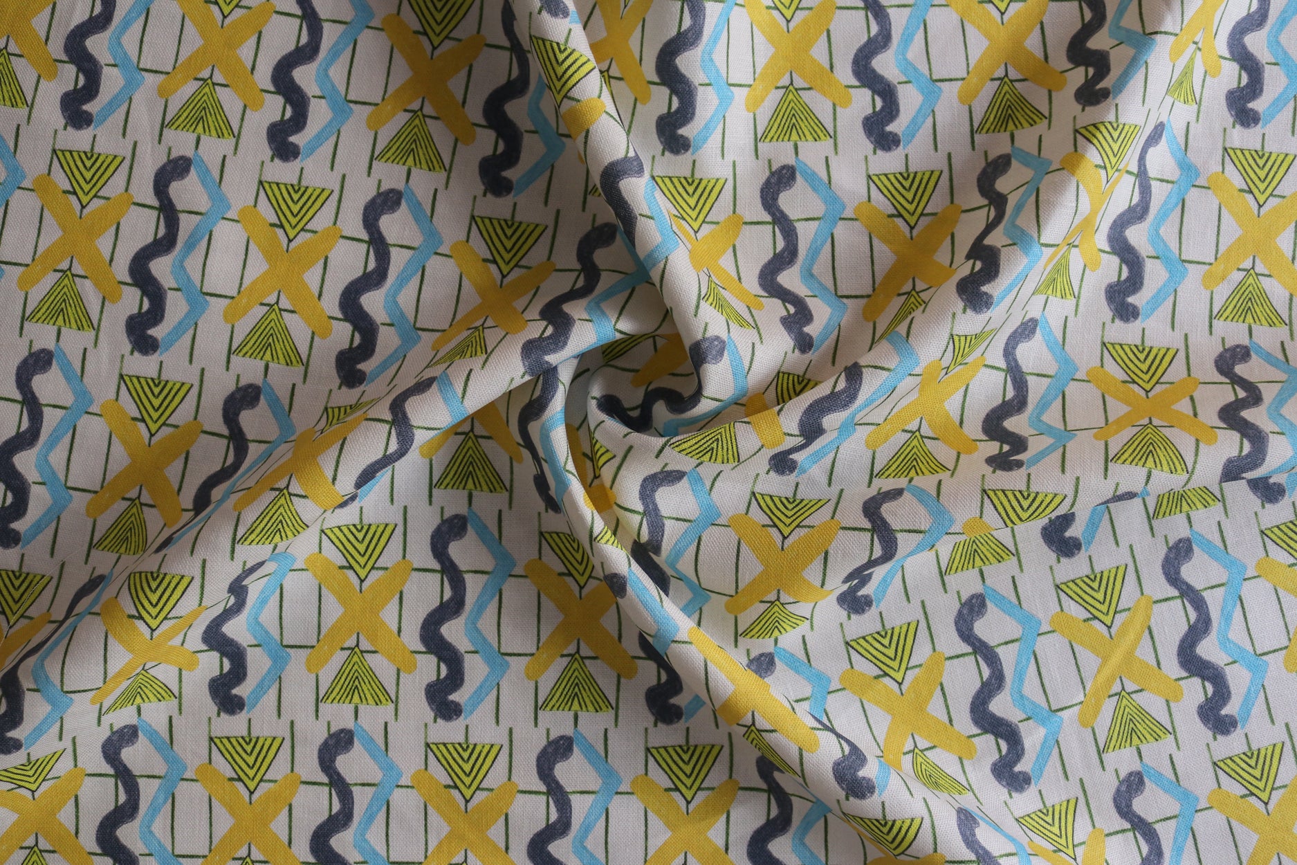 Close up of gently draped printed fabric with blue squiggles, yellow triangles,  yellow X marks on a broken grid in green on a white background. 