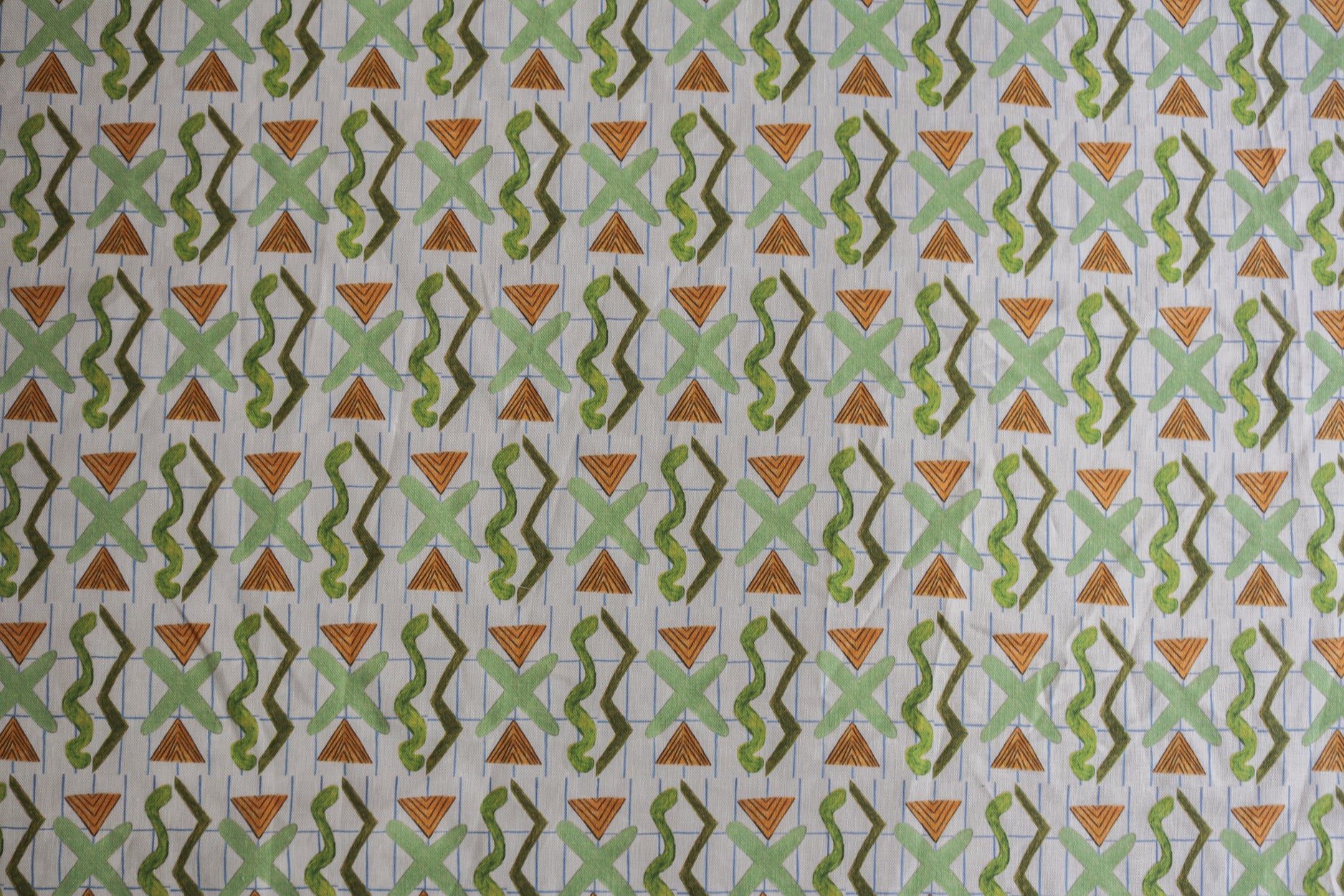 Close up of printed fabric with green squiggles, orange striped triangles, light green X marks on a broken grid in blue on a white background. 