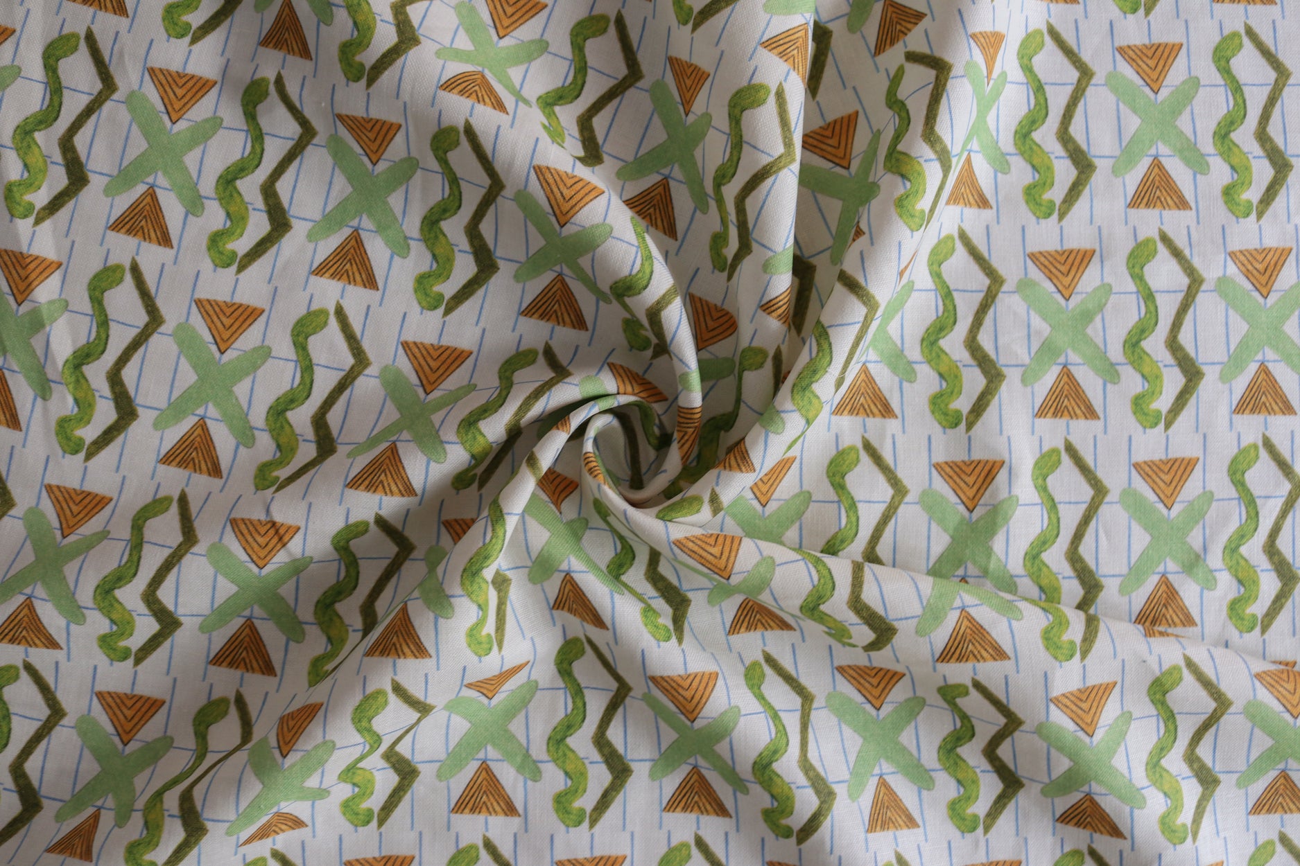 Close up of gently draped printed fabric with green squiggles, orange striped triangles, light green X marks on a broken grid in blue on a white background. 