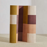 A curved partition stands on the ground, covered in an interlocking square pattern in shades of mustard, purple and pink.