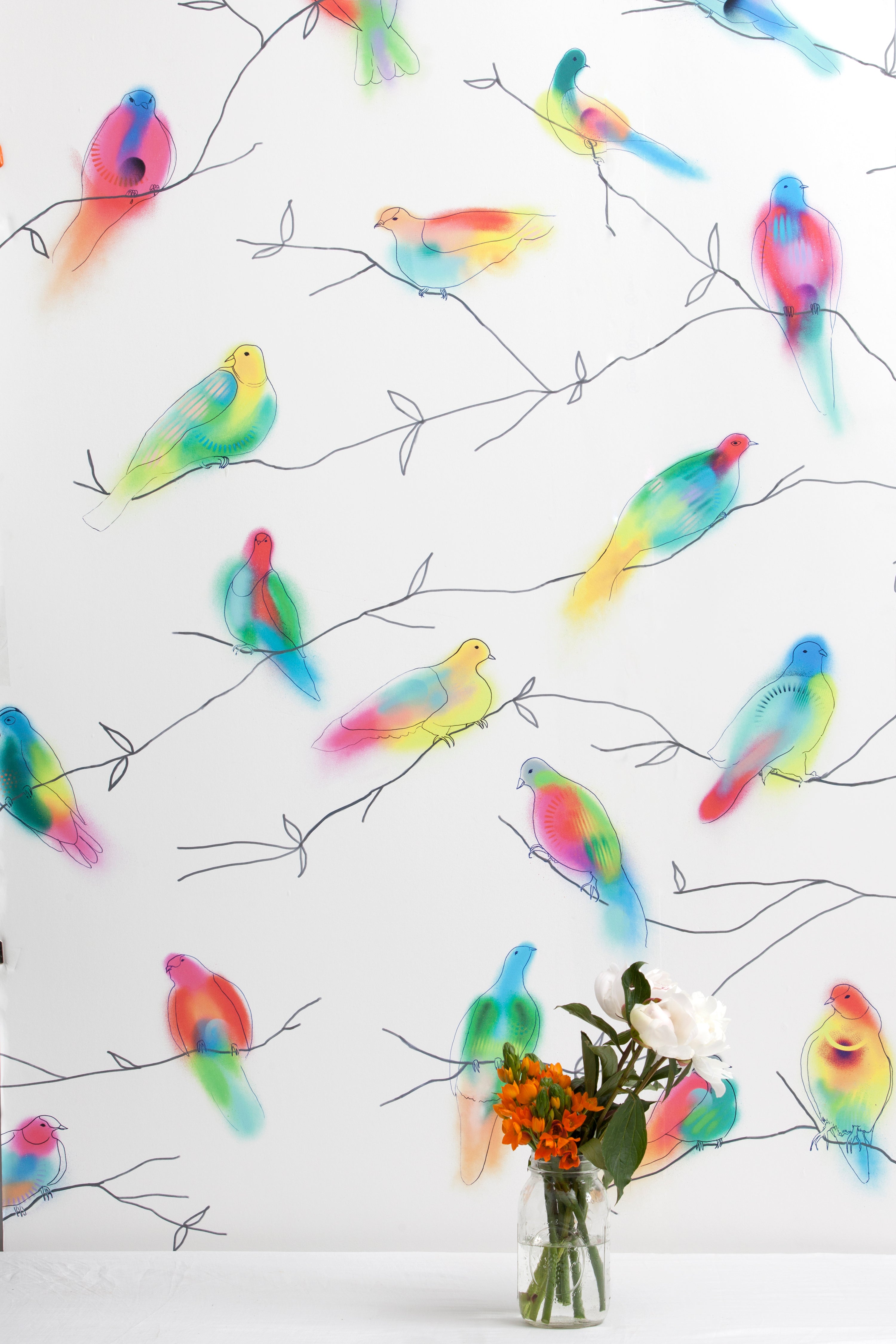 A vase of flowers stands in front of a wall papered in a painterly bird and branch print in green, yellow, pink and white.