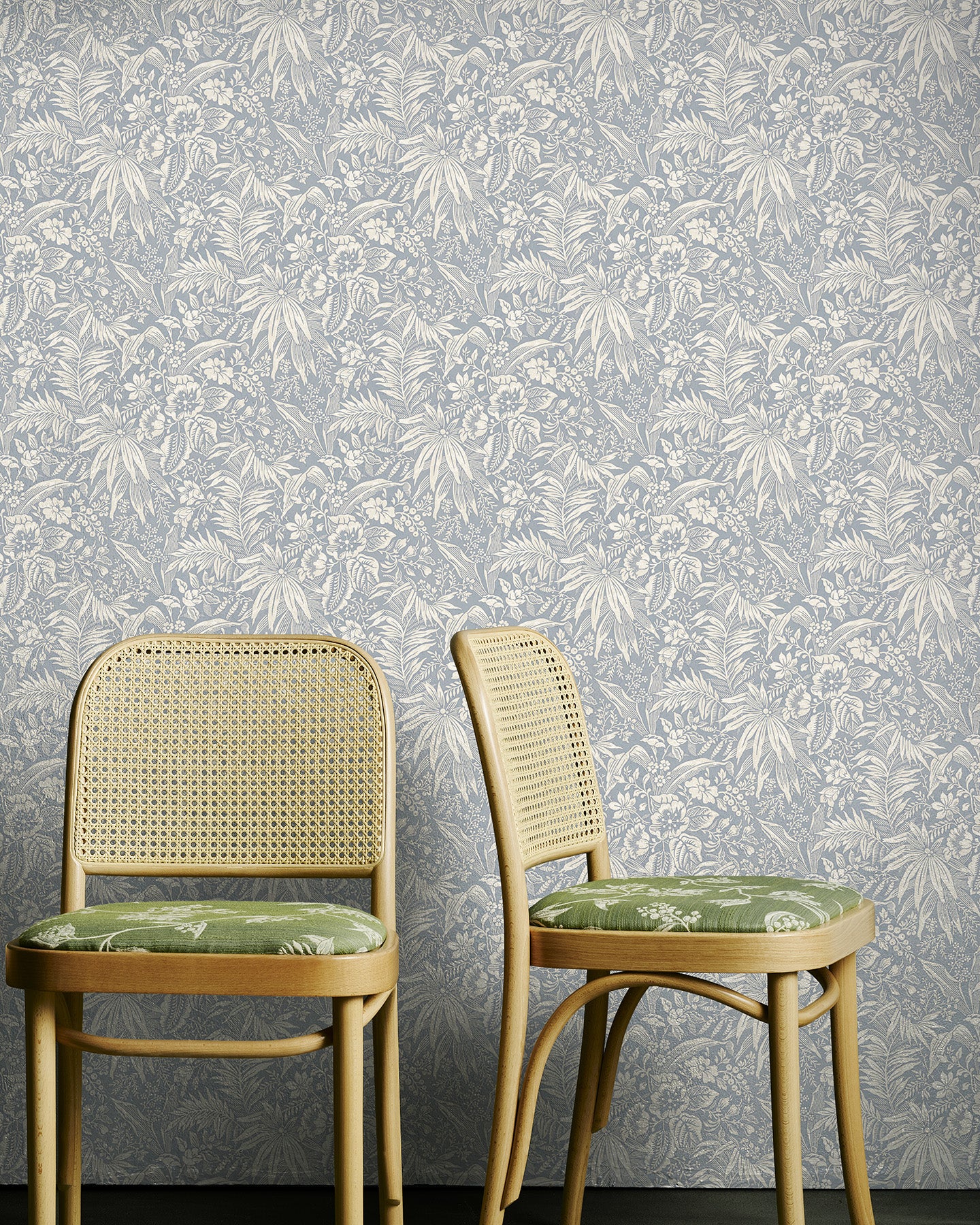 Two wooden chairs with wicker backs against a pale blue floral wallpapered wall.