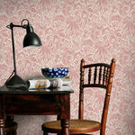 Vintage-style room with pink floral wallpaper, wooden chair, and table with lamp and newspaper.