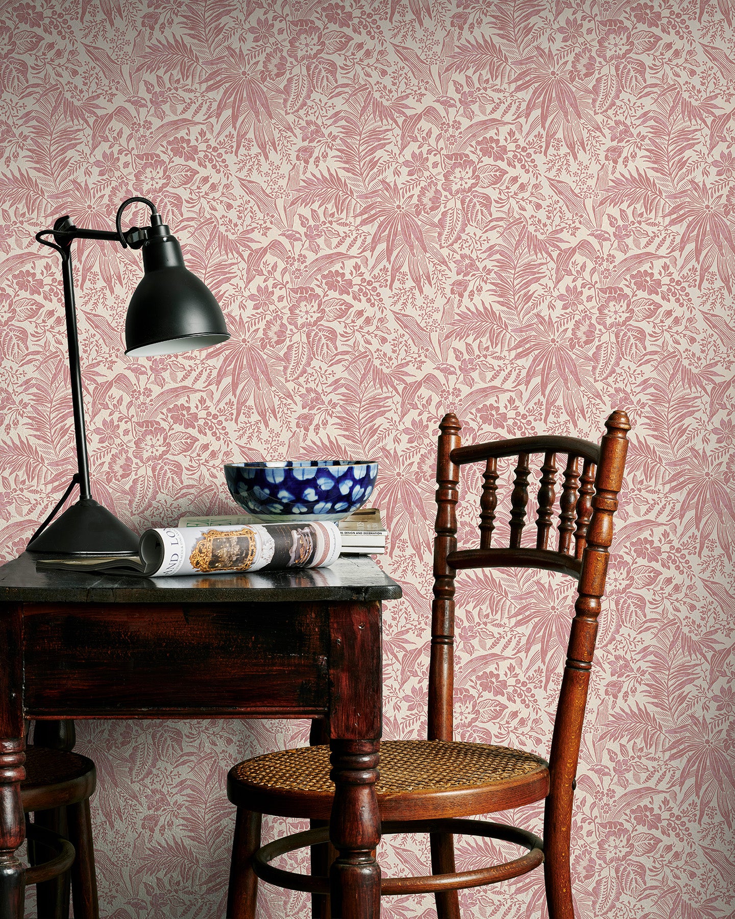 Vintage-style room with pink floral wallpaper, wooden chair, and table with lamp and newspaper.