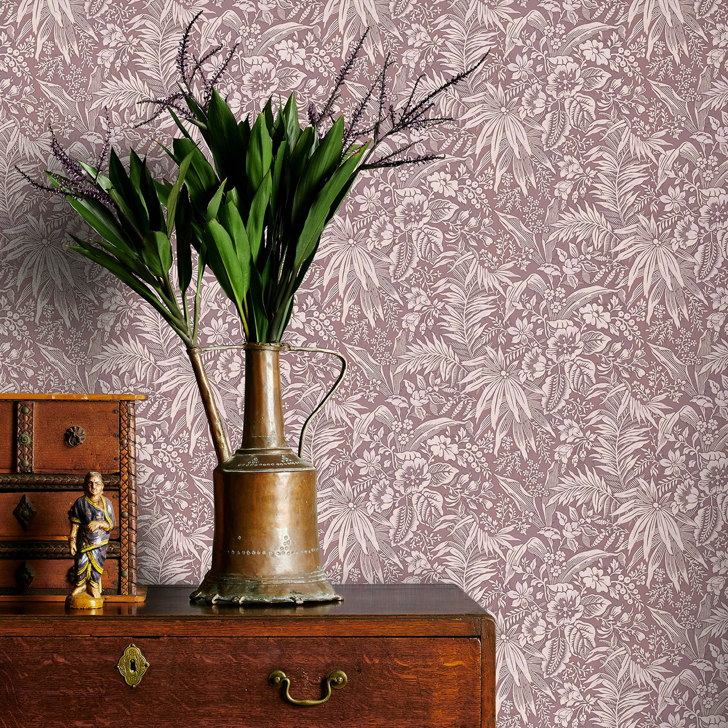 Vintage brass watering can with plants on a wooden dresser against a mauve floral wallpapered wall.