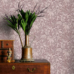 Vintage brass watering can with plants on a wooden dresser against a mauve floral wallpapered wall.