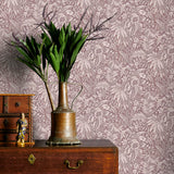 Vintage brass watering can with plants on a wooden dresser against a mauve floral wallpapered wall.