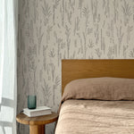 Bed with tan linens and wooden headboard, wooden side table  with white book and green glass, white floor length curtain against a black pointillist wheat frond patterned wallpaper on an ecru background. 