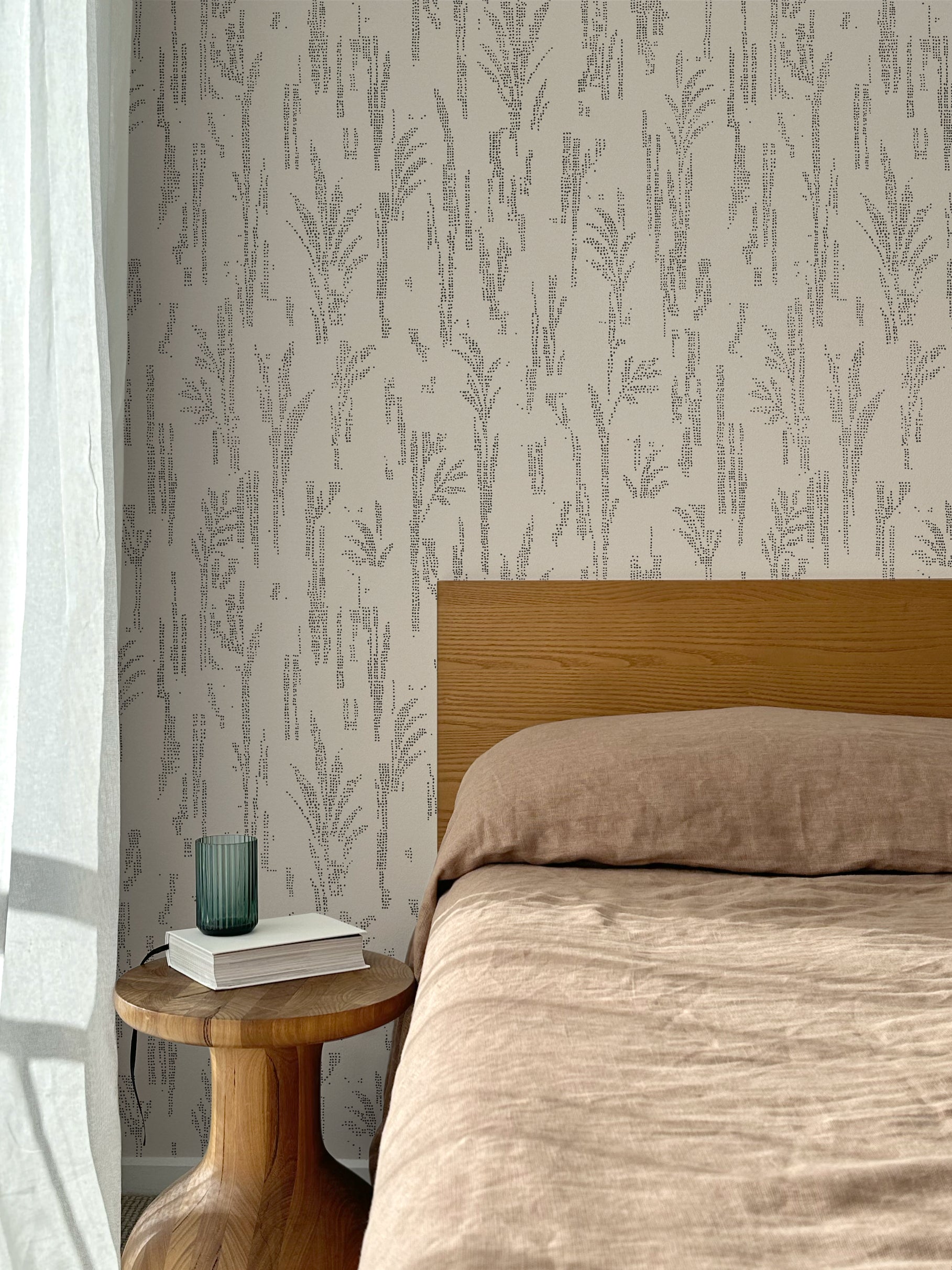 Bed with tan linens and wooden headboard, wooden side table  with white book and green glass, white floor length curtain against a black pointillist wheat frond patterned wallpaper on an ecru background. 