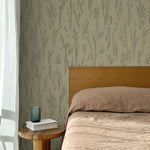 Bed with tan linens and wooden headboard, wooden side table  with white book and green glass, white floor length curtain against a black pointillist wheat frond patterned wallpaper on a sage green background. 