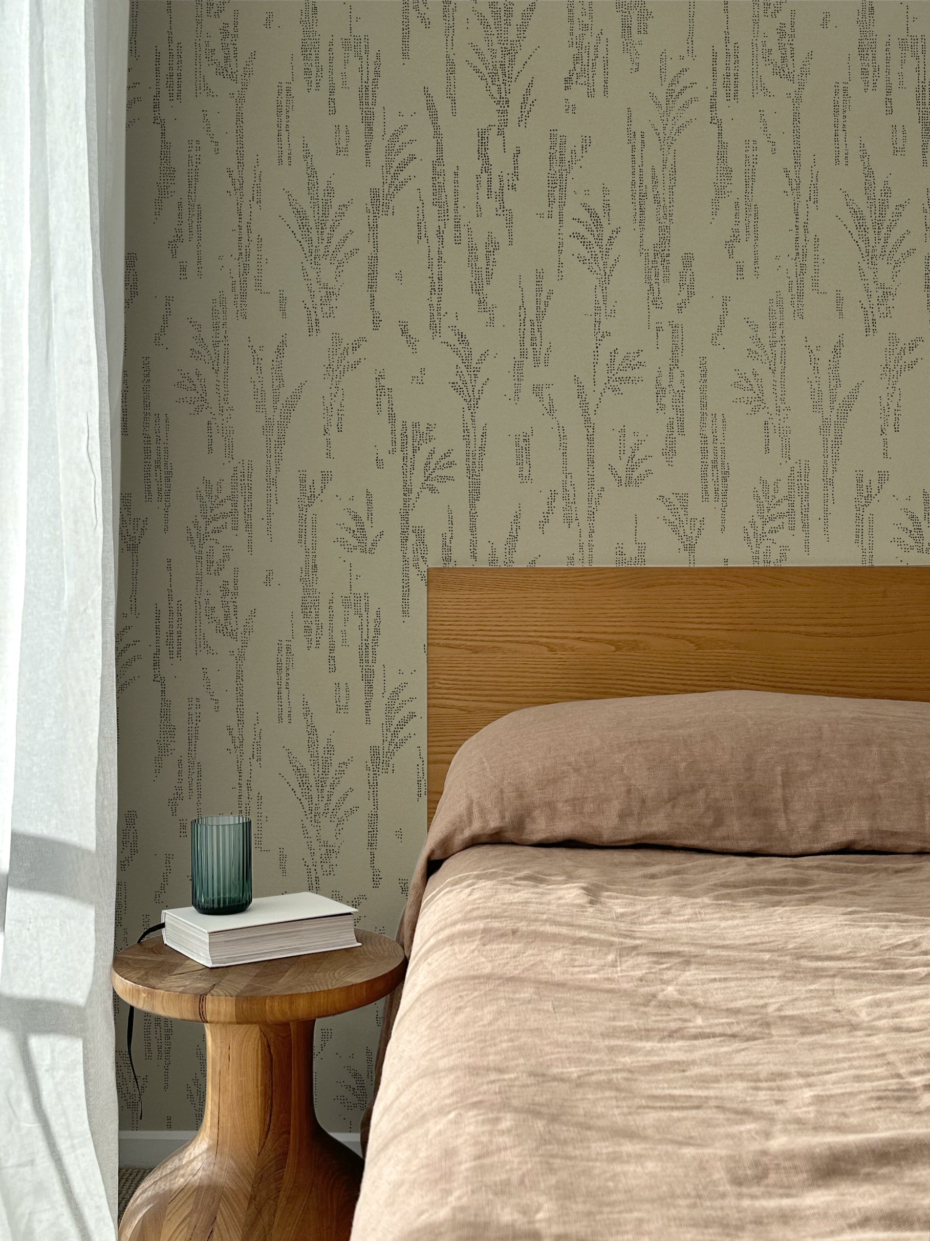 Bed with tan linens and wooden headboard, wooden side table  with white book and green glass, white floor length curtain against a black pointillist wheat frond patterned wallpaper on a sage green background. 