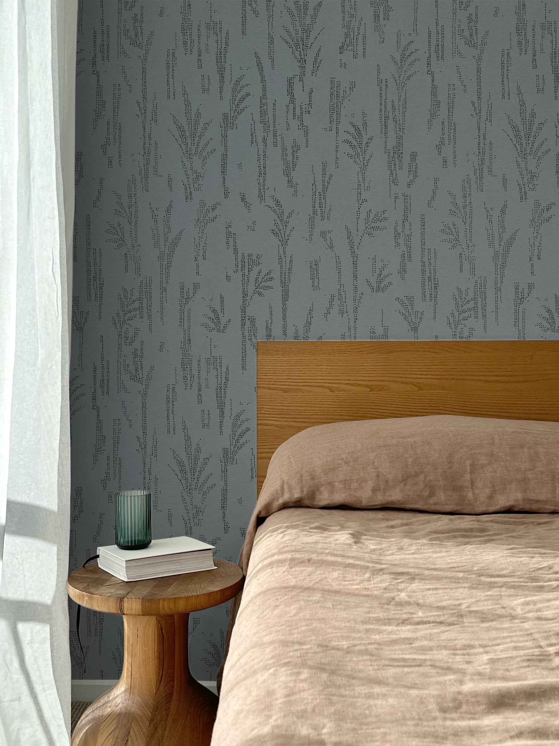 Bed with tan linens and wooden headboard, wooden side table  with white book and green glass, white floor length curtain against a black pointillist wheat frond patterned wallpaper on a steel blue-grey background. 