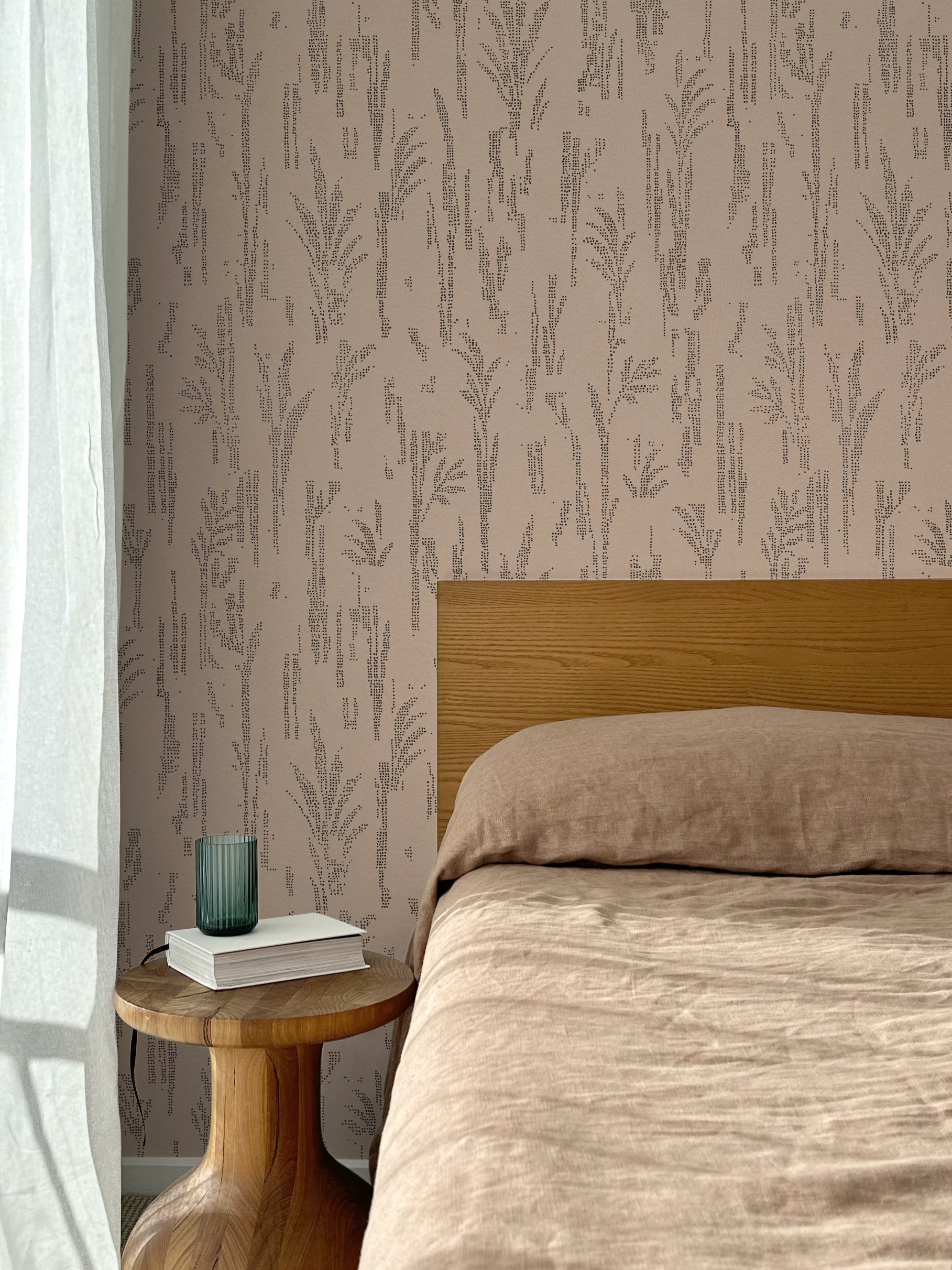 Bed with tan linens and wooden headboard, wooden side table  with white book and green glass, white floor length curtain against a black pointillist wheat frond patterned wallpaper on a soft clay pink background.