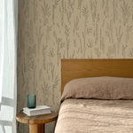 Bed with tan linens and wooden headboard, wooden side table  with white book and green glass, white floor length curtain against a black pointillist wheat frond patterned wallpaper on a tan background. 