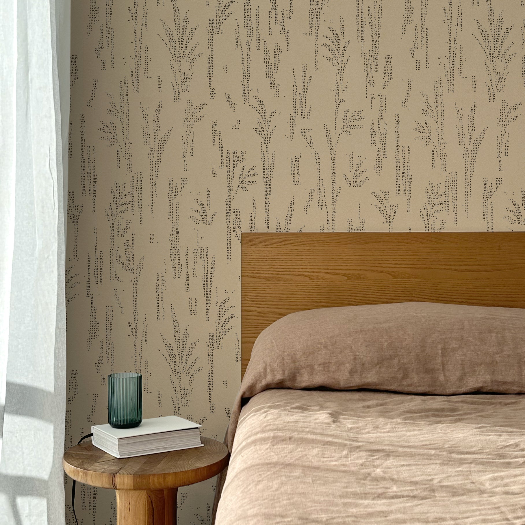 Bed with tan linens and wooden headboard, wooden side table  with white book and green glass, white floor length curtain against a black pointillist wheat frond patterned wallpaper on a tan background. 