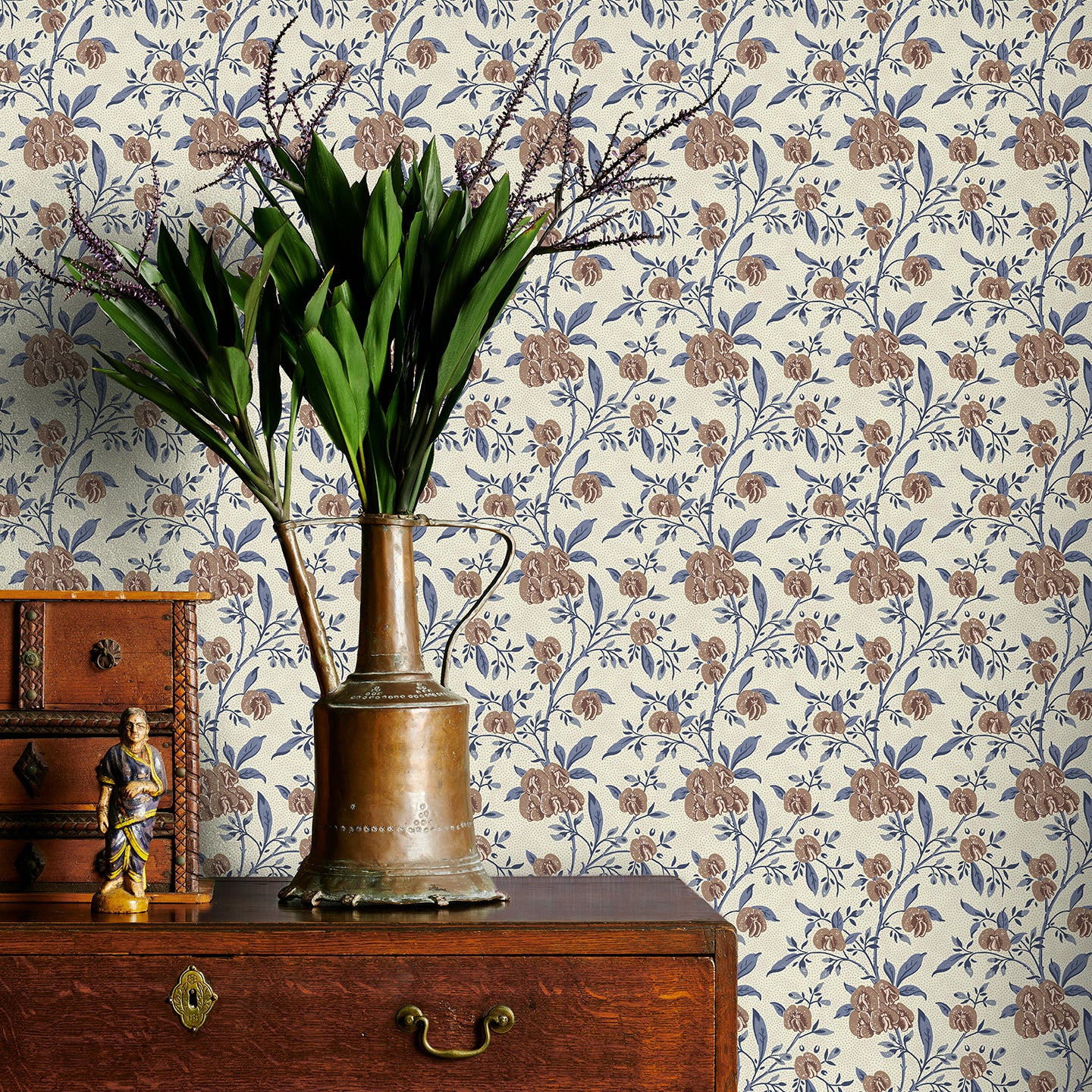 Decorative wallpaper with brown and blue floral pattern, wooden dresser with brass vase and plant, and small statue.