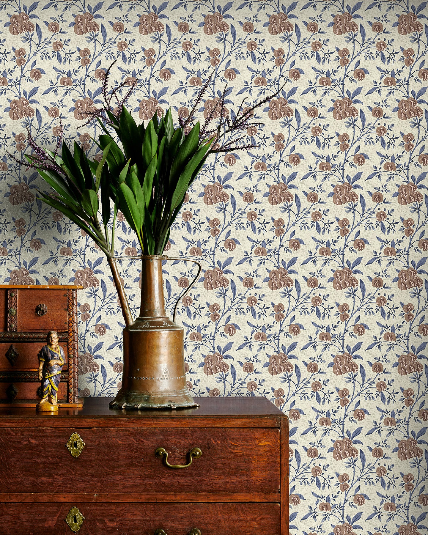 Decorative wallpaper with brown and blue floral pattern, wooden dresser with brass vase and plant, and small statue.