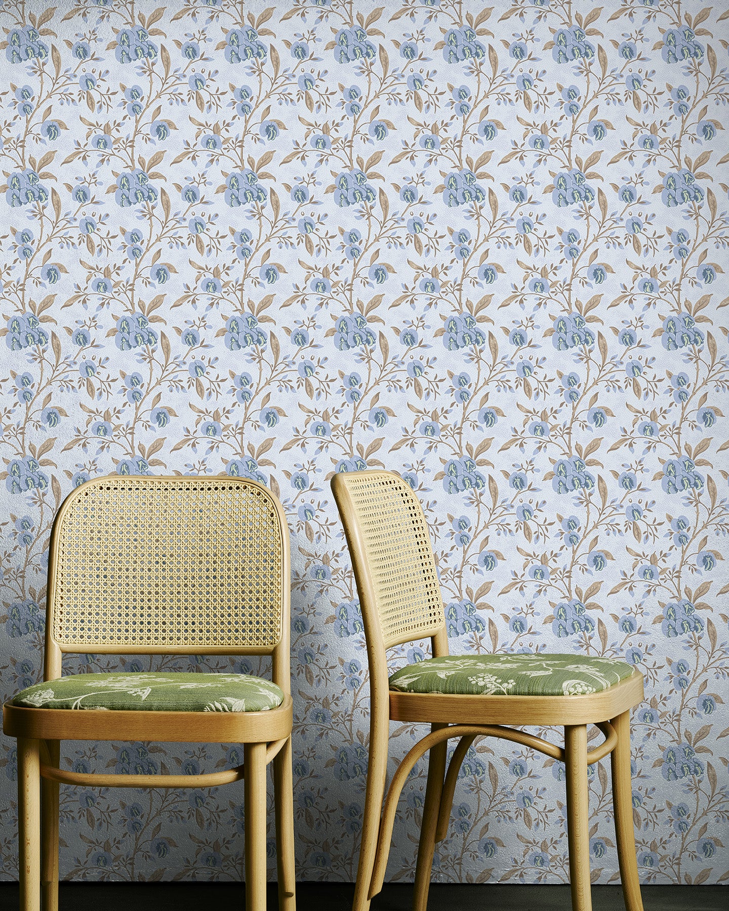 Two wicker chairs with green cushions against a blue and brown floral wallpapered wall.