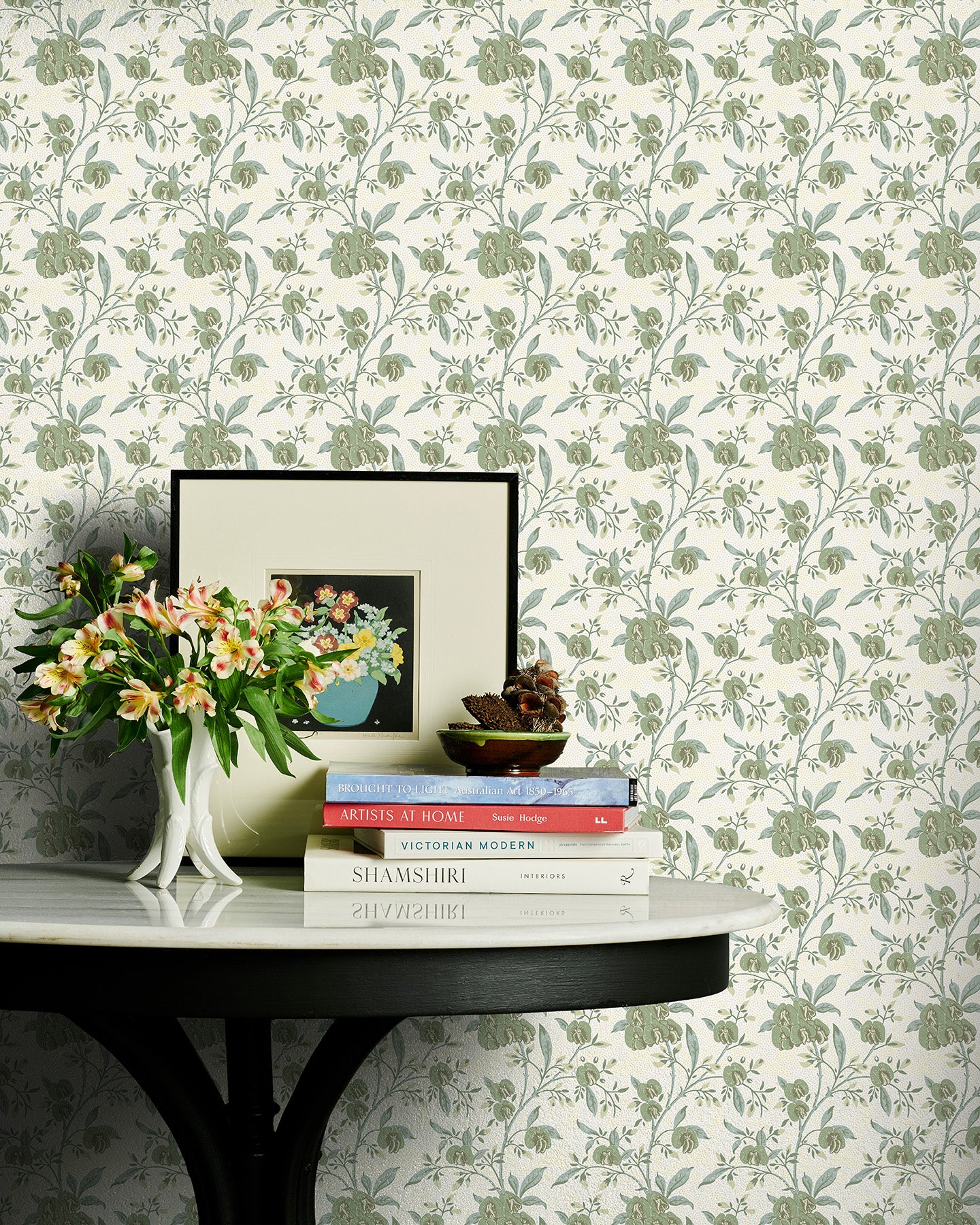 Decorative table with books and a vase against a sage green floral wallpapered wall.