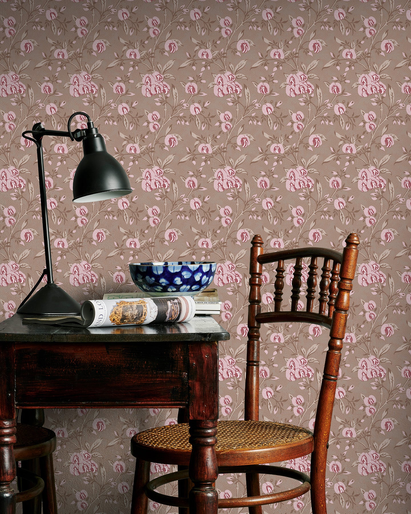 Vintage-style room with brown and pink floral wallpaper, wooden chair, and table with lamp and books.