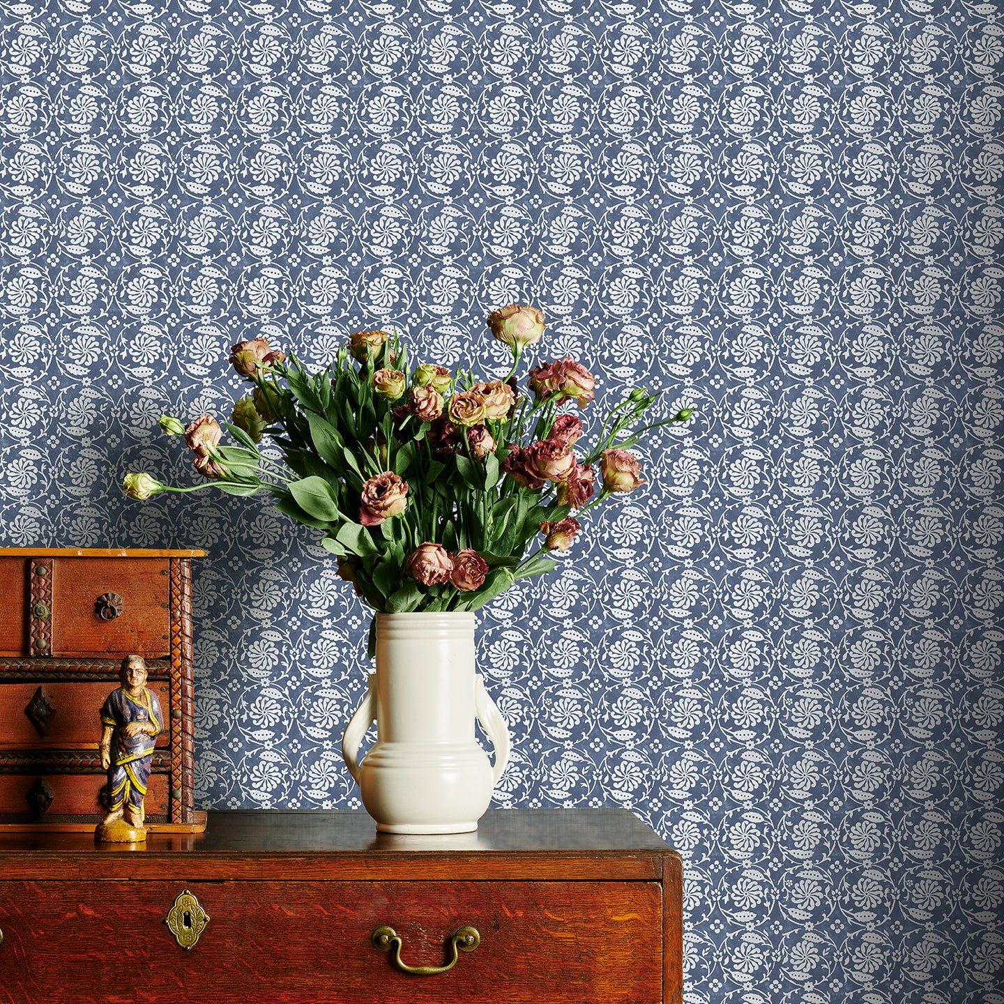 Vintage wooden dresser with a vase of flowers against a blue floral patterned wallpaper.