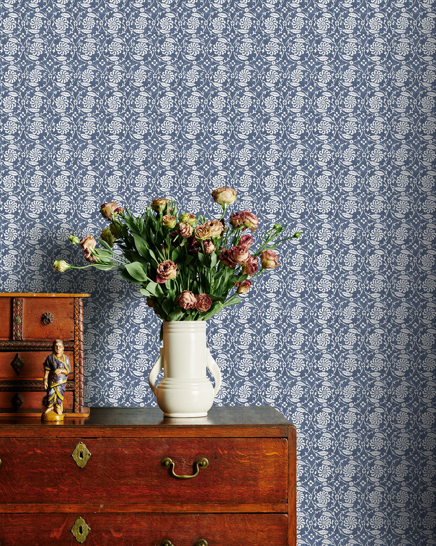 Vintage wooden dresser with a vase of flowers against a blue floral patterned wallpaper.