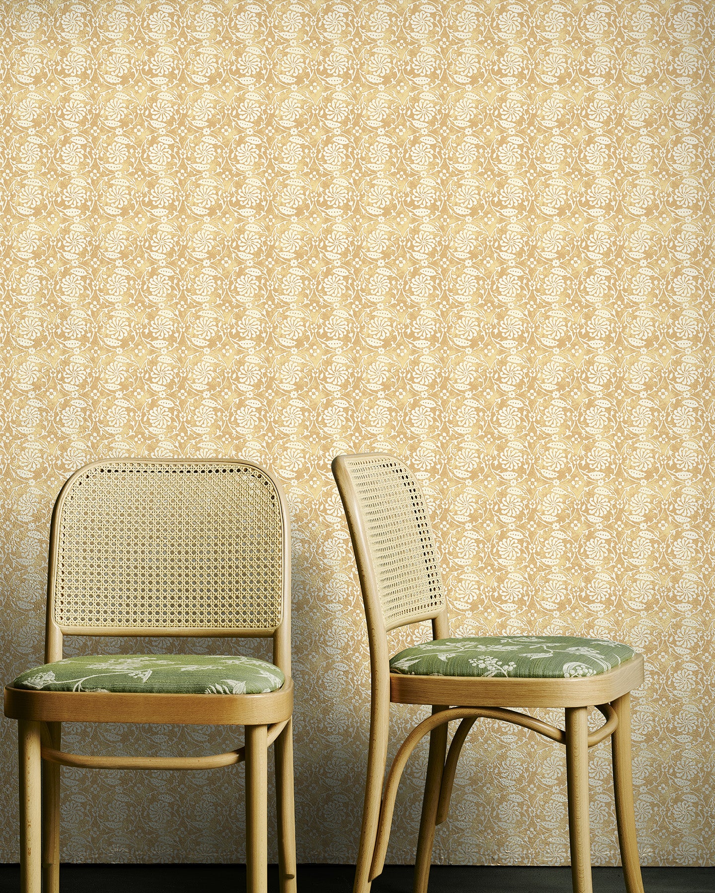 Two wooden chairs with green cushions against a yellow floral wallpapered wall.