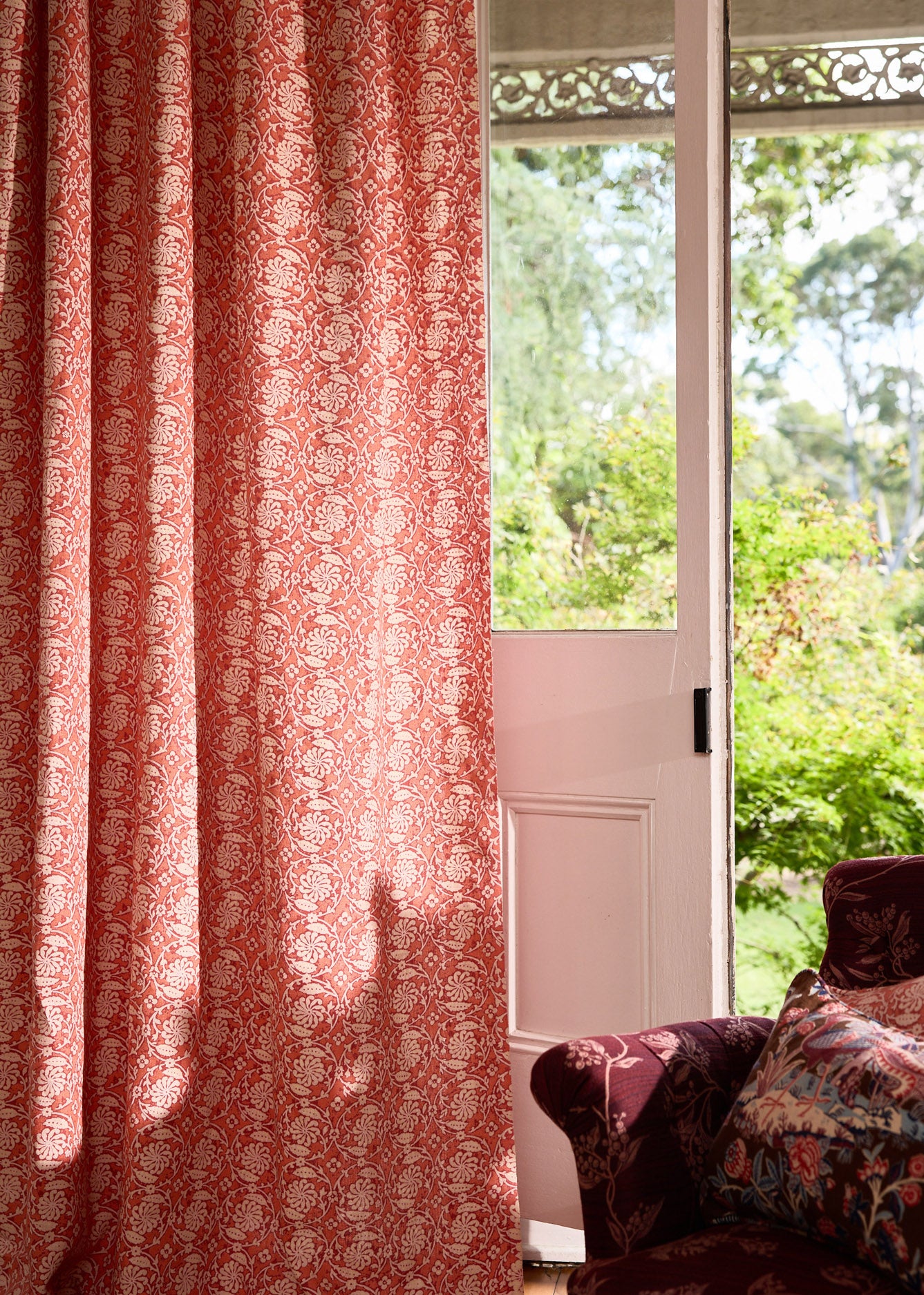 Upholstered red chair with pink patterned fabric curtain against an open door leading to the garden