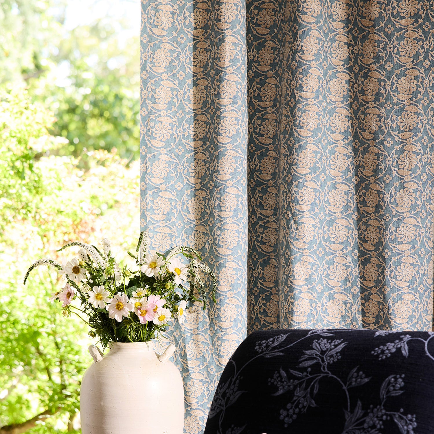 Decorative curtain with floral pattern next to a chair and vase with flowers by a window.