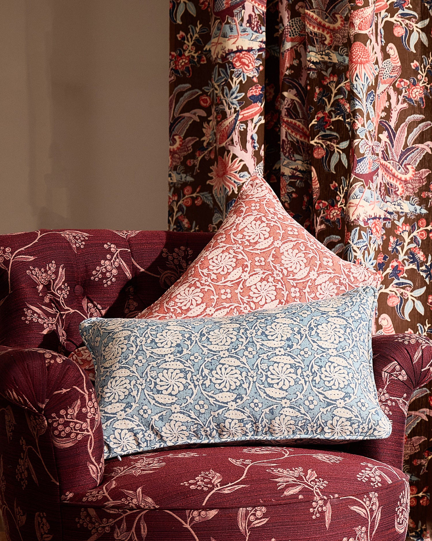 Decorative pillows on a floral-patterned armchair with floral-fauna printed curtains.
